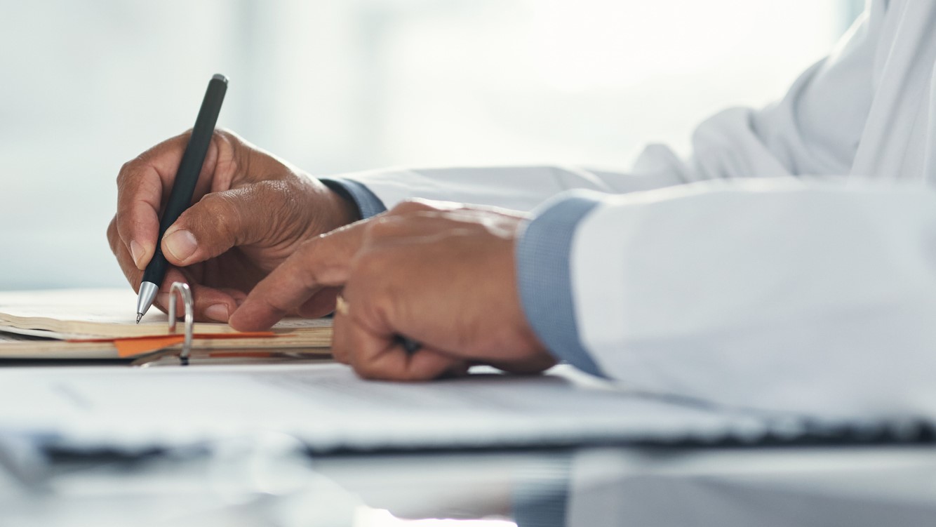 Doctor hands writing on paper or document at a desk in the hospital. Healthcare professional drafting a medical insurance letter, legal paperwork or form. A GP filing a document in a clinic office