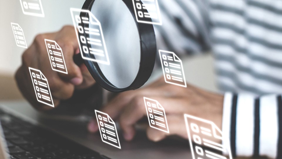 A young man sitting at his desk with a laptop on his desk, he is using a magnifying glass to look at the document file icon.