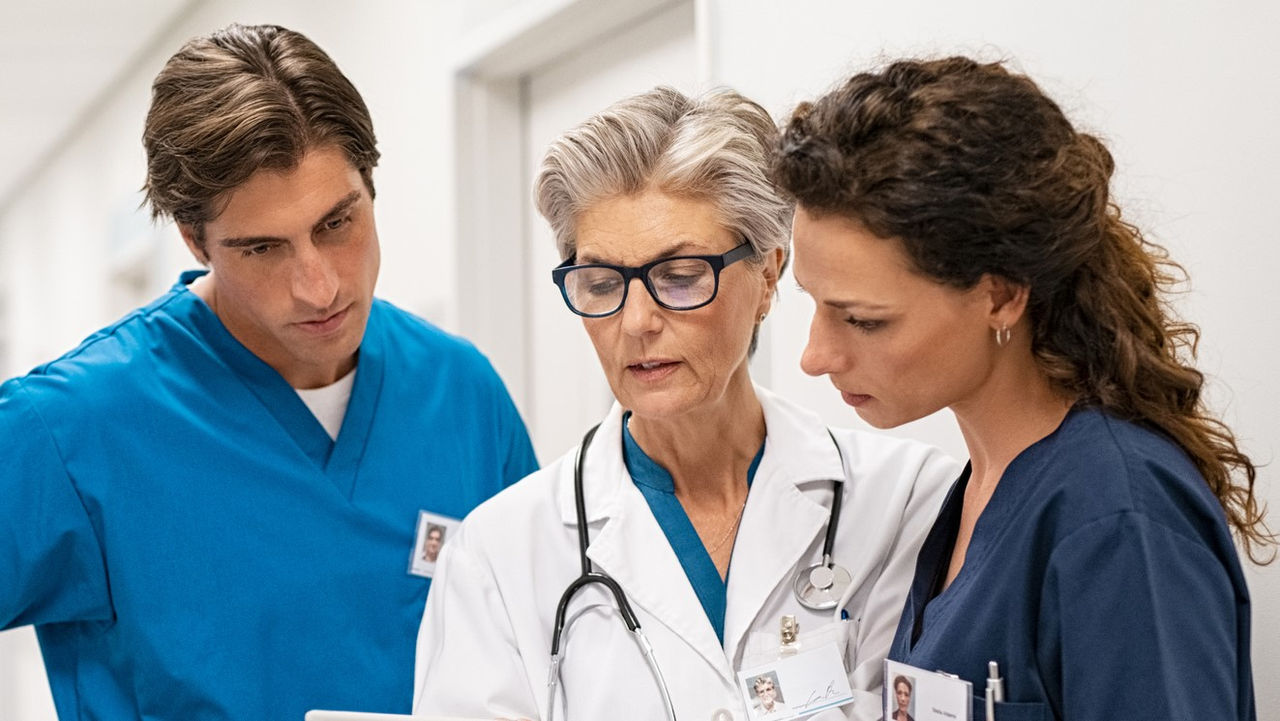 Mature female doctor discussing medical report with nurses in hospital hallway. Senior general practitioner discussing patient case status with group of medical staff after surgery. Doctor working on digital tablet while in conversation with healthcare workers, copy space.